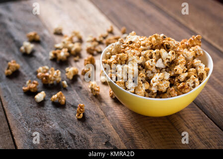 Karamell Popcorn in einem gelben Schüssel auf einem Holztisch Hintergrund. Popcorn in eine Schüssel geben und auf den Tisch verstreut Stockfoto