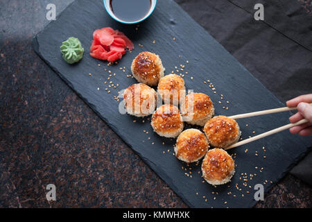Japanische Küche. Gebackene Sushi mit Krabben Fleisch und Käse auf einer Steinplatte auf dem dunklem Marmor Hintergrund. Weibliche hand Picks sticks gebacken. Ansicht von oben w Stockfoto