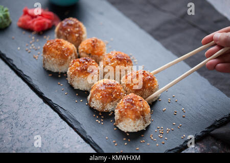 Japanische Küche. Gebackene Sushi mit Krabben Fleisch und Käse auf einer Steinplatte auf dem dunklem Marmor Hintergrund. Weibliche hand Picks sticks gebacken. Ansicht von oben. Stockfoto