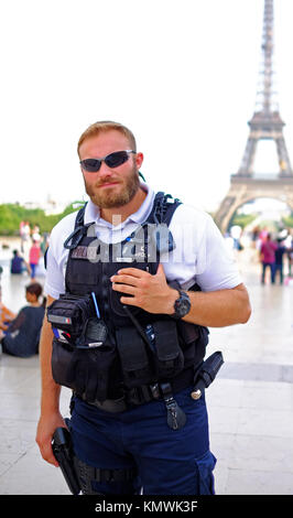 Ein bewaffneter Polizist bleibt wachsam, während sein Foto gemacht wird, seine Hand auf sein Gewehr, während auf Patrouille unter Touristen am Eiffelturm in Paris. Stockfoto