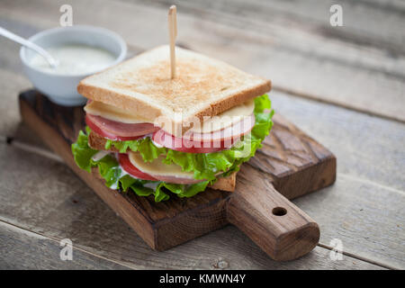 Close-up big Sandwich mit Schinken, Käse, Tomaten, Salat und weißer Soße auf geröstetem Brot auf einem rustikalen Holztisch. Stockfoto