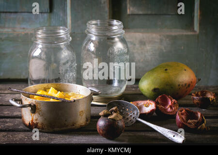 Die Mango Chutney oder Stau im Vintage Aluminium Schwenk über alte Tabelle mit leeren Glas, Mango und Mangosteen. Dunkle, rustikale Atmosphäre Stockfoto