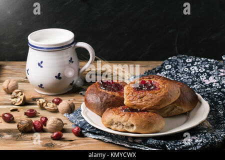 Teller mit hausgemachten Brötchen mit Marmelade, serviert auf alten Holztisch mit Nüssen, Beeren und Tasse Milch Stockfoto