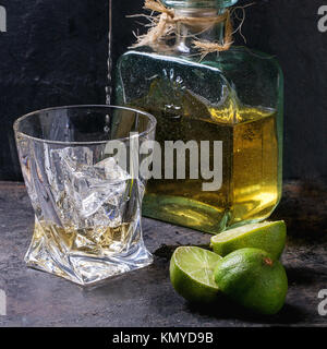 Tequila ergossen sich in Glas mit Eiswürfeln und eine Flasche Tequila, serviert mit in Scheiben geschnittenen Limetten über schwarzen Hintergrund. Quadratisches Bild Stockfoto