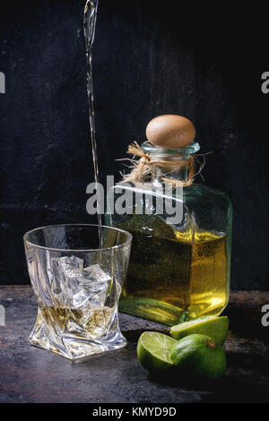 Tequila in Glas mit Eiswürfeln und Flasche Tequila, serviert mit in Scheiben geschnittenen Limetten über schwarzen Hintergrund gegossen Stockfoto