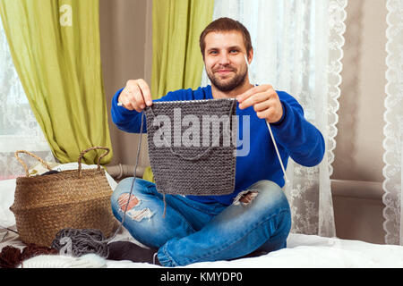 Ein junger dunkelhaariger Mann in einem blauen Pullover und Jeans zeigt, wie er ein Stück von einem grauen pullover mit Stricknadeln aus natürlichen Strings gebunden und setzt sich auf Stockfoto