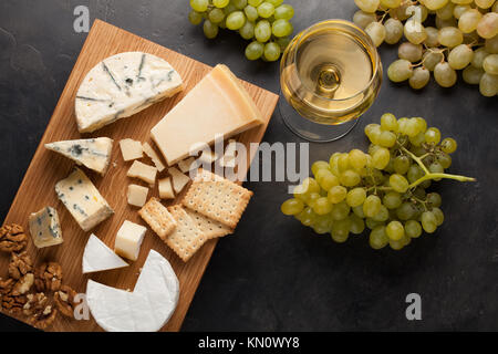 Verschiedene Käsesorten mit weißen Trauben, Walnüsse, Cracker und weißen Wein auf einem Holzbrett. Essen für ein romantisches Date auf einem dunklen Stein Hintergrund. Ansicht von oben. Stockfoto