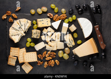 Verschiedene Käsesorten mit weißen Trauben, Walnüsse, Cracker und weißen Wein auf einem Stein. Essen für ein romantisches Date auf einem dunklen Hintergrund. Ansicht von oben. Stockfoto