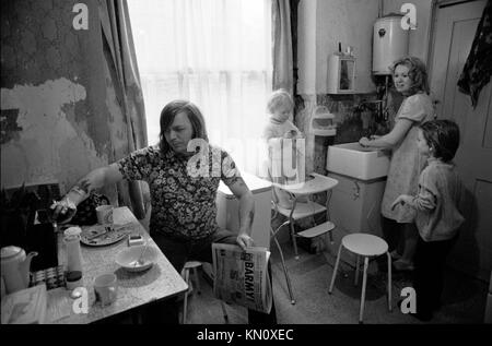 1970er Slum-Gehäuse, Küche dient auch als Badezimmer. Die Familie isst zusammen. Armut. Ascot Gas-Wasser-Boiler an der Wand über dem Butler Spüle 1972 Fulham, England 70s UK HOMER SYKES Stockfoto