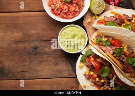 Drei Mexikanische Tacos mit Marmor, Black Angus Rindfleisch und Gemüse auf alten rustikalen Tisch. Mexikanische Gerichte mit Soßen Guacamole und Salsa in Schalen. Ansicht von oben Stockfoto