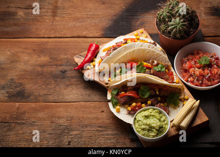 Drei Mexikanische Tacos mit Marmor, Black Angus Rindfleisch und Gemüse auf alten rustikalen Tisch. Mexikanische Gerichte mit Soßen Guacamole und Salsa in Schalen. Ansicht von oben Stockfoto