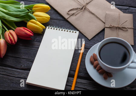 Notizblock mit Bleistift neben den Tulpen, Kaffee und Briefumschläge auf Holz- Hintergrund. Sicht von oben mit kopieren. Stockfoto