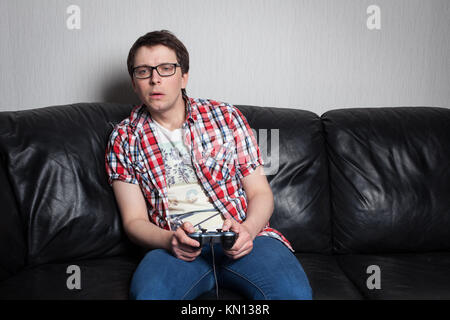 Junger Kerl mit Brille und dem roten Hemd Videospiele auf den Joystick und sitzt auf einem schwarzen Ledersofa. Stockfoto