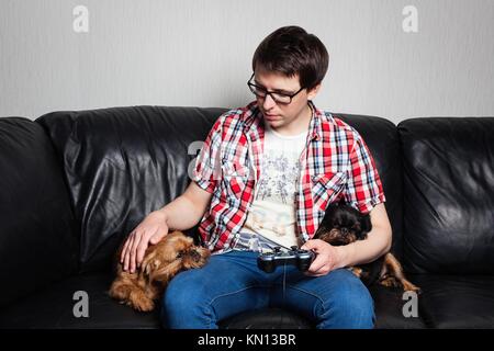 Ein junger Mann in einem roten T-Shirt und Blue Jeans sitzt zu Hause und spielt Videospiele zusammen mit ihrem Hund. Der Kerl hat eine Pause zum Haustier ihre Haustiere. Die Konz Stockfoto
