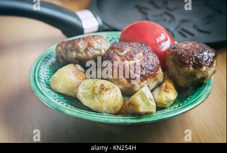 Auf dem Tisch in einem keramischen Platte der gebratenen Frikadellen und Scheiben aus gerösteten gewürzten Kartoffeln. Auf der Platte liegt neben der Tomate. Stockfoto