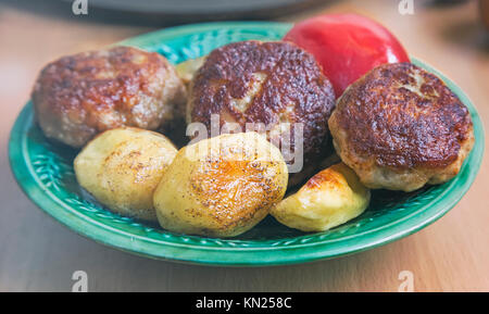 Auf dem Tisch in einem keramischen Platte der gebratenen Frikadellen und Scheiben aus gerösteten gewürzten Kartoffeln. Auf der Platte liegt neben der Tomate. Stockfoto