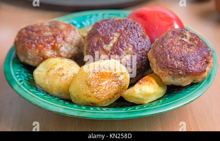 Auf dem Tisch in einem keramischen Platte der gebratenen Frikadellen und Scheiben aus gerösteten gewürzten Kartoffeln. Auf der Platte liegt neben der Tomate. Stockfoto