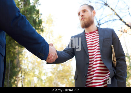 Low Angle Portrait von junge Bartgeier Geschäftsmann Händeschütteln mit unkenntlich Partner im Freien treffen im Park Stockfoto