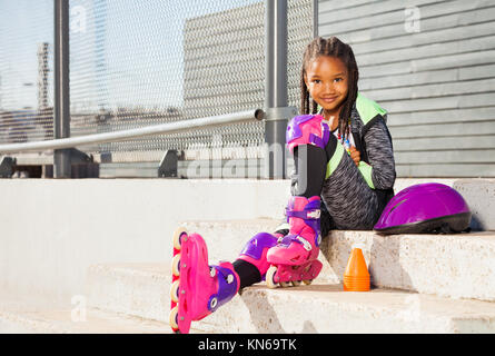 Cute afrikanisches Mädchen in roller blades auf der Treppe der Skate Park am sonnigen Tag sitzen Stockfoto