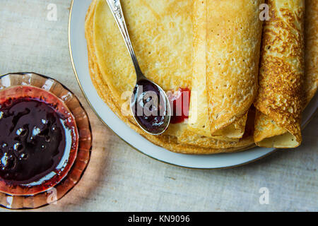 Stapel gerollt Golden Crepes auf weiße Platte schwarze Johannisbeere Konfitüre in Crystal Rosette Löffel Bettwäsche Tischdecke Draufsicht Stockfoto