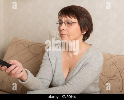 Ältere Frau in Gläsern sitzen auf der Couch mit Fernbedienung in der Hand Stockfoto