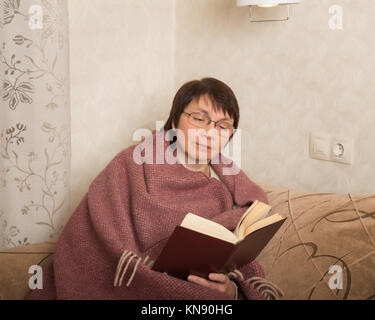 Ältere Frau sitzen auf der Couch eingehüllt in eine Decke und ein Buch lesen Stockfoto