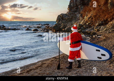 Mann in Santa Kostüm mit SUP Board auf Winter Meer Stockfoto