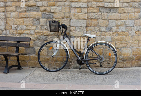 Nahaufnahme von einem Fahrrad mit einem schwarzen Metallgitter Warenkorb Gleitstift gegen eine Mauer aus Stein. Ein metall Bank ist auf der linken Seite. Stockfoto