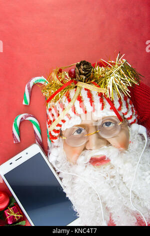 Santa mit Smartphone- und Ohrhörer, auf Rot mit Weihnachtsverzierungen Stockfoto