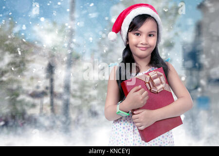 Gerne asiatische Mädchen in rot santa hut mit Weihnachtsgeschenk auf schneefall Hintergrund Stockfoto