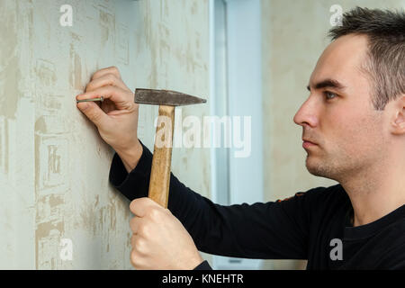 Hübscher junger Mann mit Hammer zu Hause. Kerl, Renovierung zu Hause stehend vor der Wand. Ein Mann Hammer einen Nagel in Gips an der Wand unter der Tapete Stockfoto