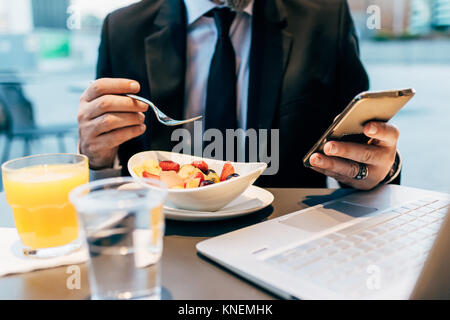 Reifen Geschäftsmann, Sitzen, Draußen, Essen, Frühstück, über Smartphone, Laptop auf dem Tisch, mittlere Partie Stockfoto
