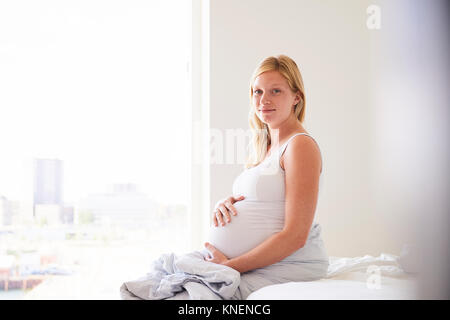 Portrait von schwangere junge Frau sitzt auf dem Bett mit den Händen auf den Magen Stockfoto