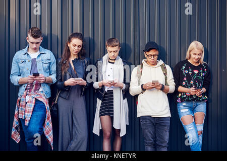 Reihe von fünf jungen Erwachsenen Freunde gegen schwarze Wand lehnt an Smartphones suchen Stockfoto
