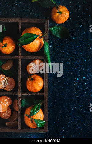 Mandarinen in einer Holzkiste auf einem dunklen Hintergrund. Ein Sortiment von Clementinen mit grünen Blättern. Dunkle essen Fotografie mit leuchtendem orange Obst und Stockfoto
