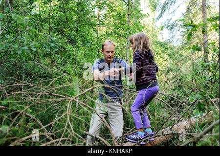 Überwinden von Hindernissen beim Gehen in den Wald. Die Erwachsenen hilft den Mädchen, den gefallenen Baum zu überqueren. Stockfoto