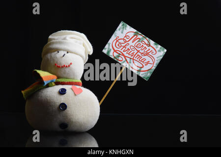 Schneemann Abbildung eine Pappe auf einem Stick mit Text "Frohe Weihnachten!" geschrieben, und mit Tannenzweigen umgeben. Low-key-Fotografie. Stockfoto