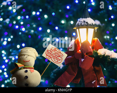 Schneemann Abbildung Holding ein Plakat. Text "Frohe Weihnachten!" darauf geschrieben. Straßenlaterne in Red Ribbon und Tannenzweigen gewickelt. Schönes Bokeh. Stockfoto