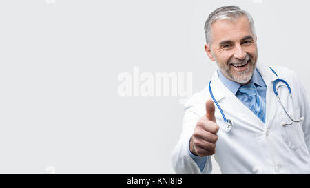 Sicher ein Lächeln auf den Lippen reife Arzt posieren und einen Daumen nach oben Stockfoto