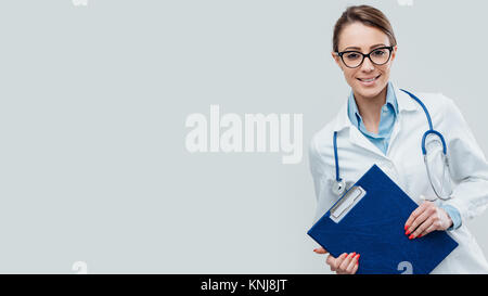 Selbstbewussten jungen Frau Doktor Lächeln und posieren mit Klemmbrett und Stethoskop Stockfoto