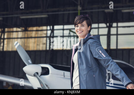 Lächelnd, fröhlich weibliche pilot posiert im Hangar mit Arme wie Flügel, Propeller Flugzeug auf dem Hintergrund, Reisen und Luftfahrt-Konzept Stockfoto