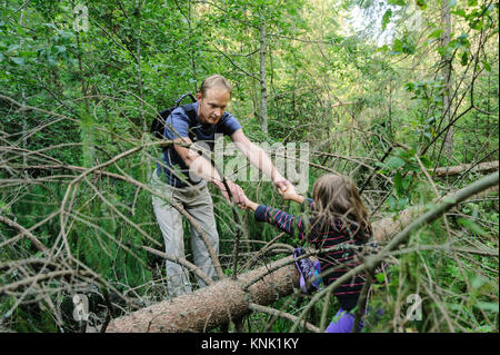 Überwinden von Hindernissen beim Gehen in den Wald. Die Erwachsenen hilft den Mädchen, den gefallenen Baum zu überqueren. Stockfoto