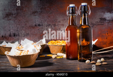 Verschiedene Bier Snacks in Platten wie Pistazien, kleinen Brezeln und Erdnüsse in der Nähe von zwei Bierflaschen auf dunklem Holzschreibtisch. Nahrungsmittel und Getränke Konzept Stockfoto