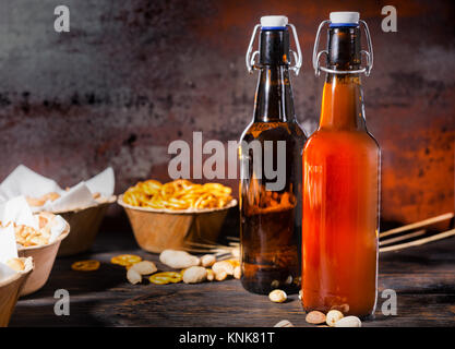 Verschiedene Bier Snacks in Platten wie Pistazien, kleinen Brezeln und Erdnüsse in der Nähe von zwei Flaschen gefilterte und ungefilterte Bier auf dunklem Holzschreibtisch. Essen Stockfoto