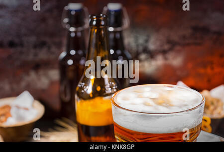 Kopf des Bieres Schaum im Glas in der Nähe von Flaschen und Teller mit Pistazien, kleinen Brezeln, Erdnüsse auf dunklem Holzschreibtisch. Nahrungsmittel und Getränke Konzept Stockfoto