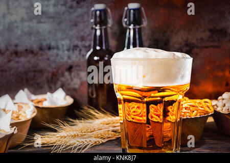 Glas mit frisch gezapften Bier und großem Kopf, der Schaum in der Nähe von Flaschen, Weizen und die Platten mit den Snacks auf dunklem Holzschreibtisch. Nahrungsmittel und Getränke Konzept Stockfoto