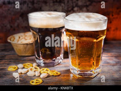 Zwei große Gläser mit frisch gegossen und dunklen Bier in der Nähe verstreut kleine Brezeln und Pistazien auf dunklem Holzschreibtisch. Nahrungsmittel und Getränke Konzept Stockfoto