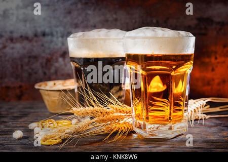 Zwei große Gläser mit frisch gezapften Bier in der Nähe von dunklen und hellen Weizen, verstreuten kleinen Brezeln und Pistazien auf dunklem Holzschreibtisch. Nahrungsmittel und Getränke co Stockfoto