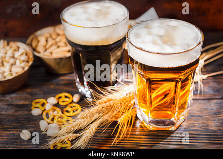 Blick von oben auf die zwei große Gläser mit frisch gezapften Bier in der Nähe von dunklen und hellen Weizen, verstreuten kleinen Brezeln und Pistazien auf dunklem Holzschreibtisch. Essen und Stockfoto