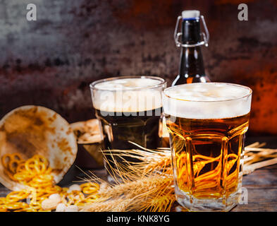 Zwei große Gläser mit frisch gezapften Bier in der Nähe von dunklen und hellen Weizen, Bier Flasche und verstreuten kleinen Brezeln auf dunklem Holzschreibtisch. Speisen und Getränke c Stockfoto